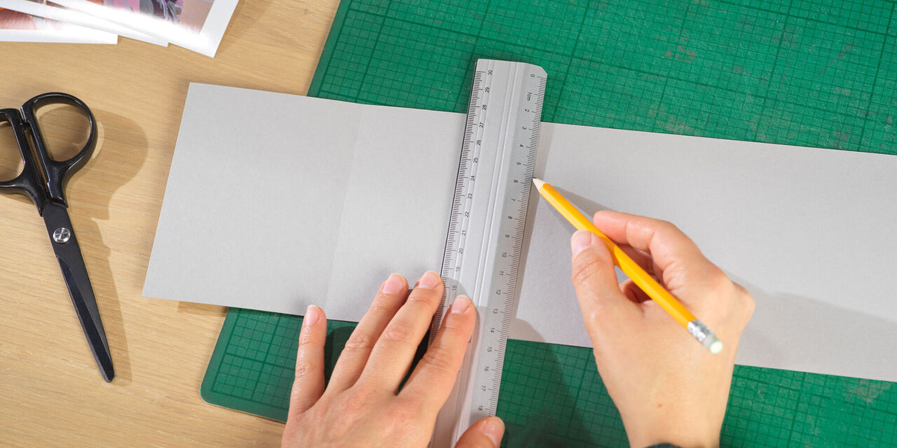Două mâini folosesc creioane și liniar pentru a trasa linii pentru fotografii pe o foaie de carton gri.