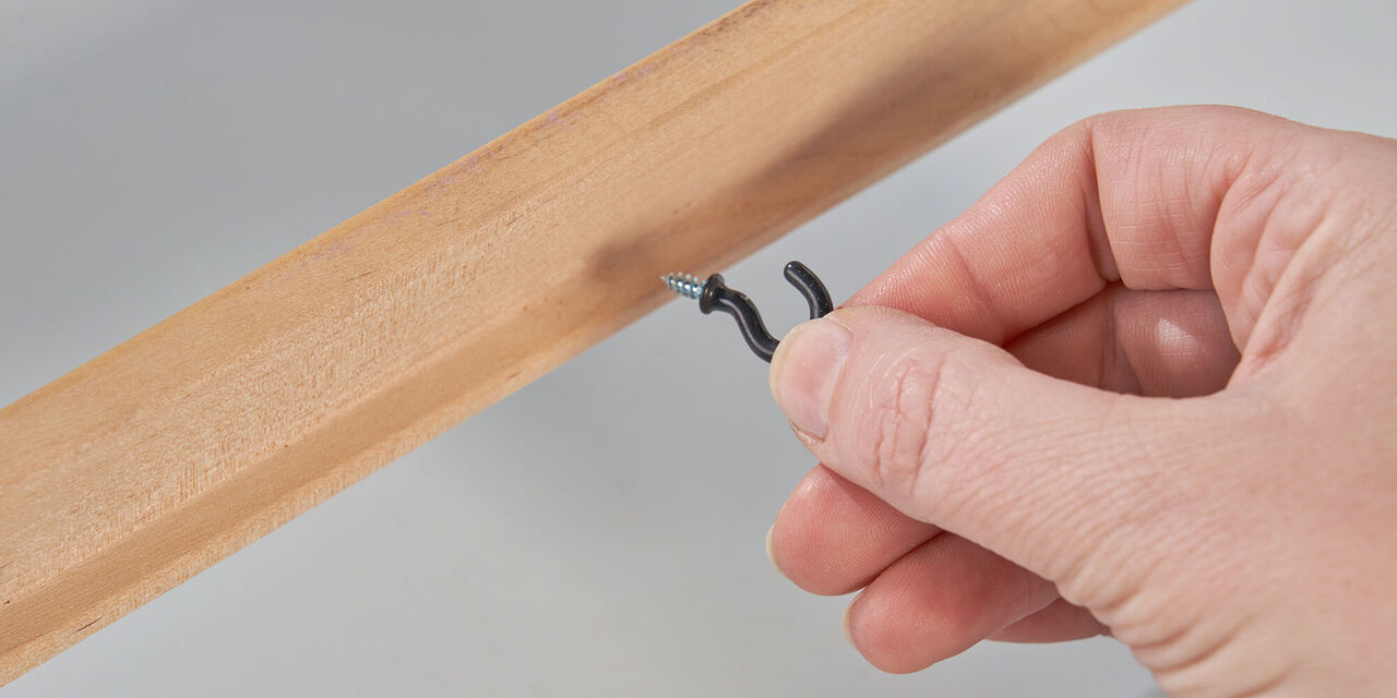 Prim-planul unei mâini care înșurubează un cârlig mic și răsucit într-un cadru de lemn.
