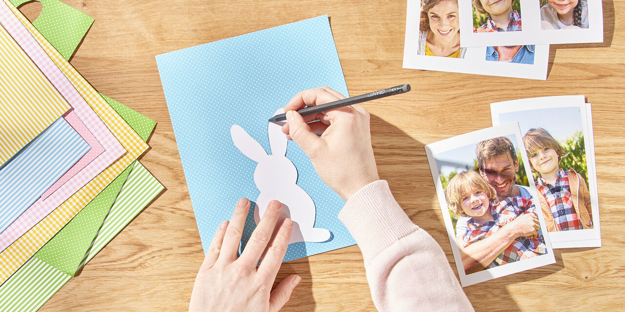 Două mâini desenează un iepuraș de Paște pe hârtie folosind șablonul. Lângă el sunt  fotografii instant ale familiei.