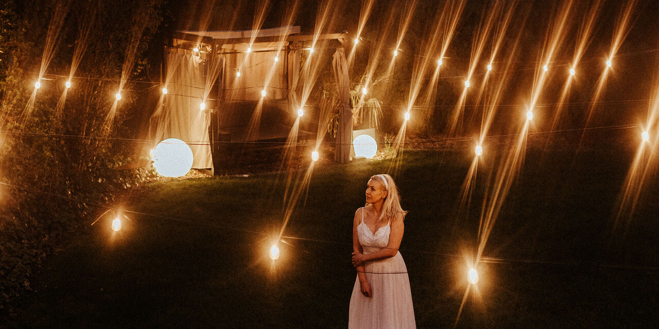 O femeie într-o rochie albă sub cerul nopții. Ea este înconjurată de o lumină ambientală, cu un pavilion luminat în fundal.