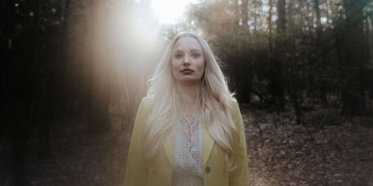 Doamnișoara blondă în geacă galbenă care stă în pădure este surprinsă în lumina soarelui din spate, fundalul fiind ușor estompat.