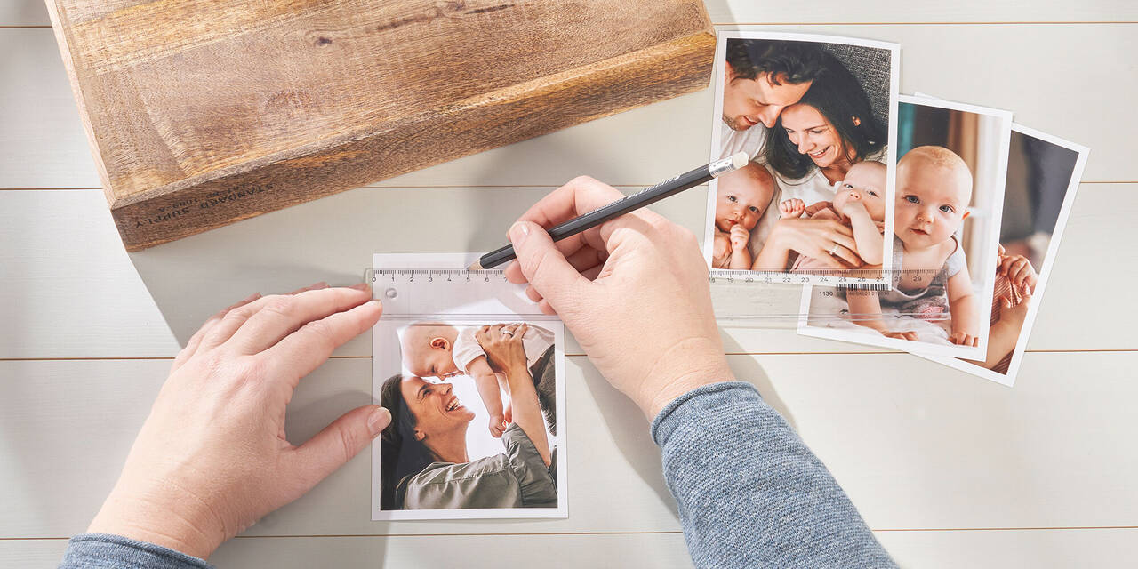 Două mâini care desenează un semn pe o fotografie cu un creion și o riglă. Lângă se află un bloc de lemn și alte trei fotografii.