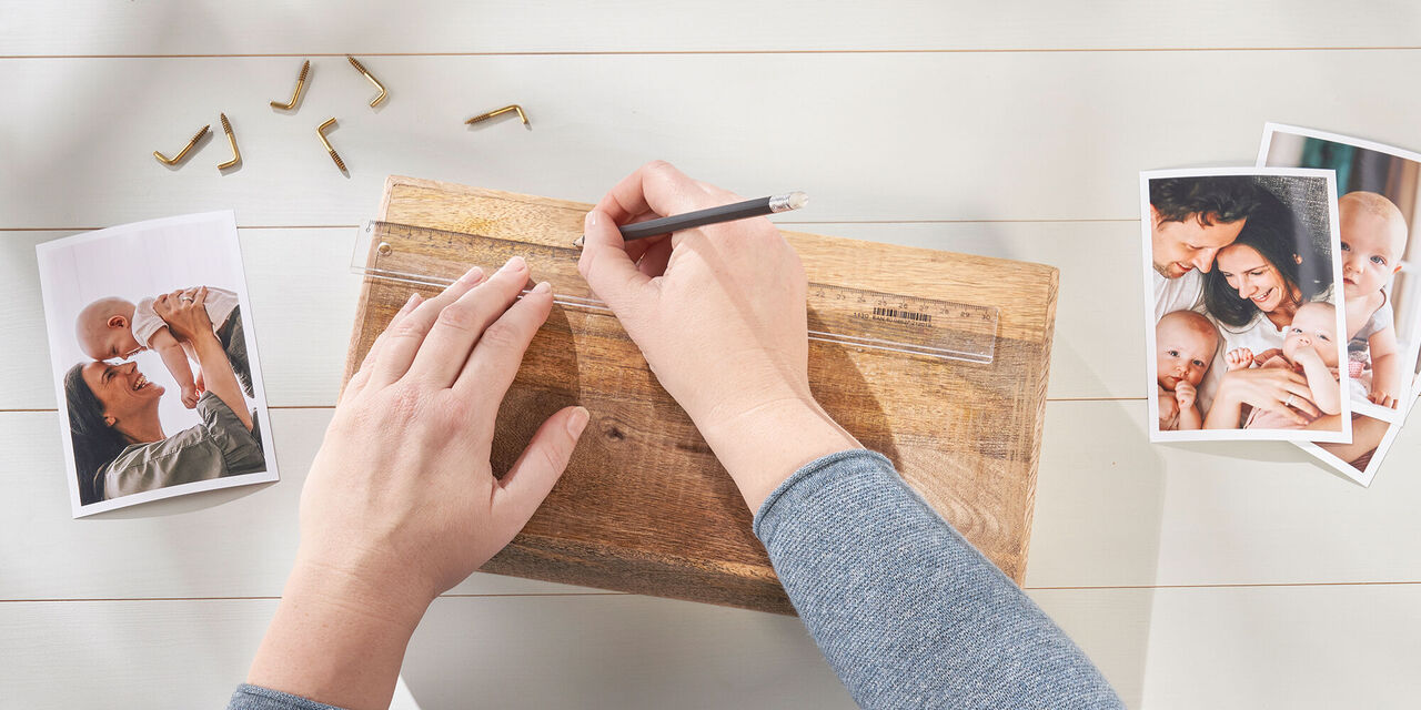 Prim-plan: două mâini care desenează semne pe un bloc de lemn cu creionul și rigla. Lângă ele se află fotografii și șuruburi cu cârlige.