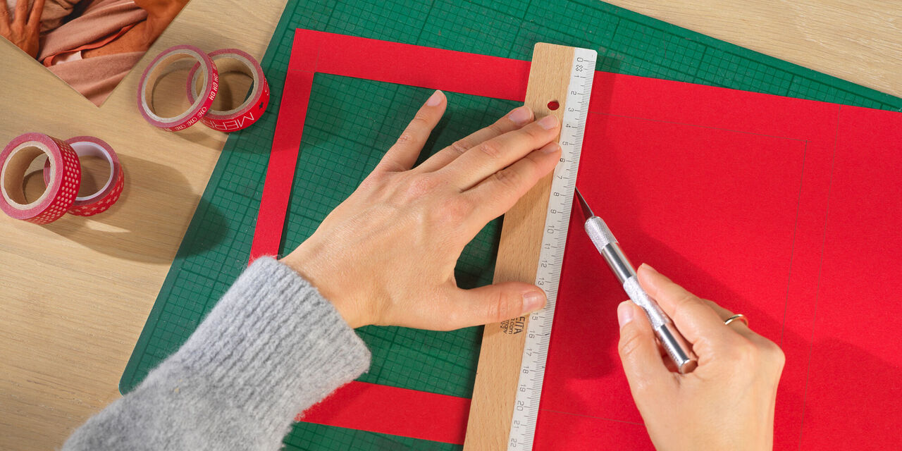 Două mâini țin un liniar și un creion. Una dintre cele patru ferestre care urmează să fie realizate este deja decupată din carton roșu.